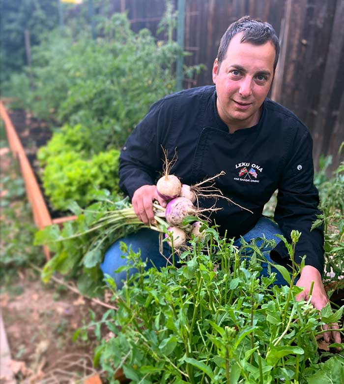 A chef from Leku Ona Restaurant Boise Idaho kneels in a garden with fresh root vegetables.