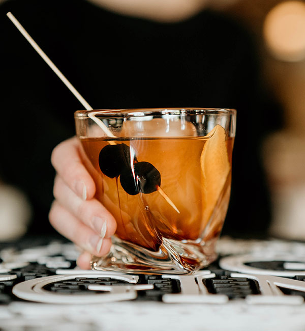 A hand holding a cocktail at Leku Ona Restaurant Boise Idaho, garnished with cherries and orange peel.